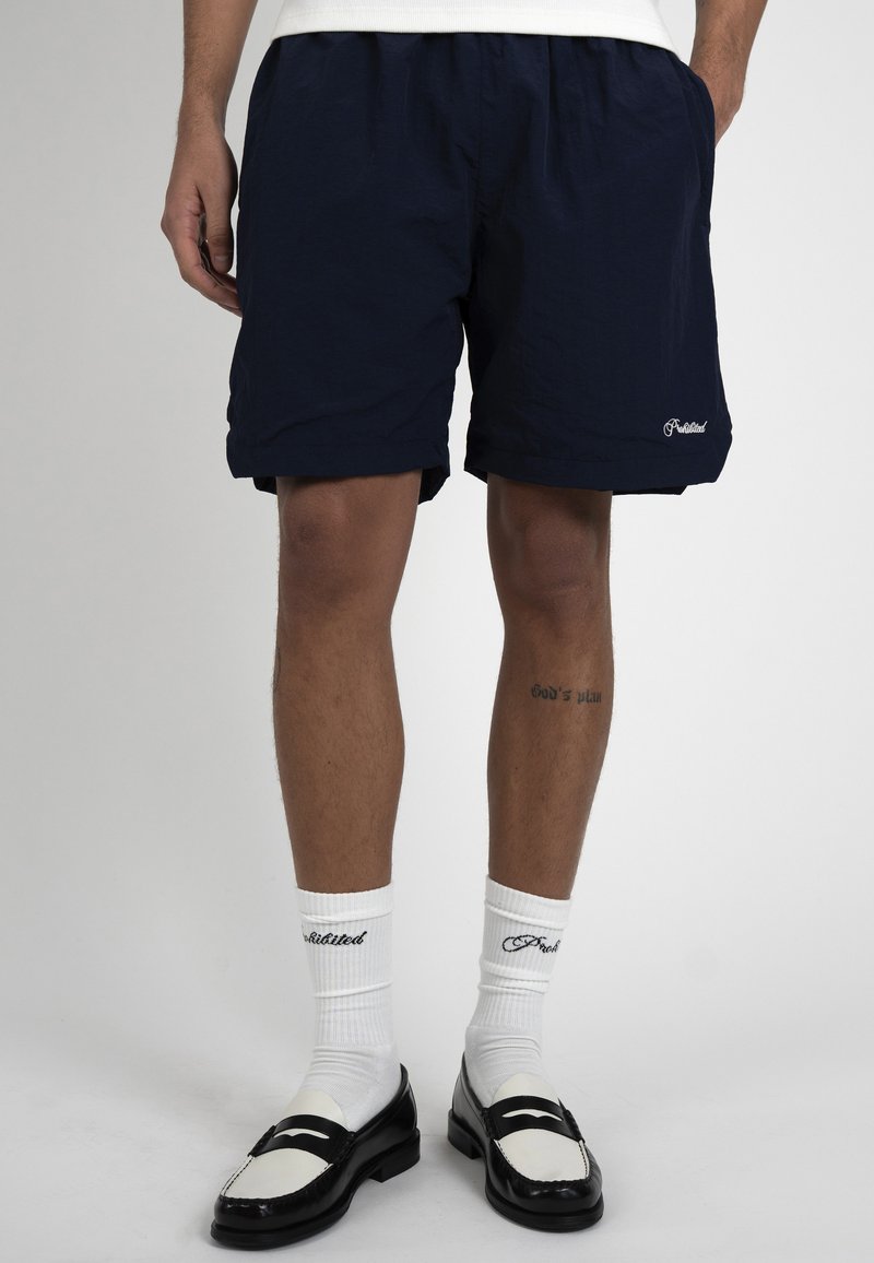Homme portant un short bleu marine, des chaussettes blanches avec texte noir, et des mocassins noir et blanc, debout sur un fond uni.