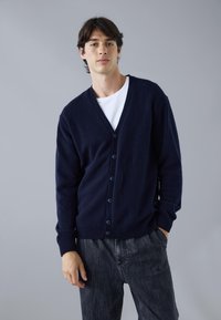 Mörkblå knappradad cardigan i ett mjukt material med V-ringning, ribbade ärmslut och en avslappnad passform, som kombineras med mörka strukturerade byxor.