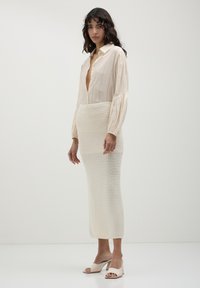 Woman with wavy dark hair wearing a beige open-collar blouse, a cream knitted midi skirt, and beige open-toe block heels standing against a plain background.