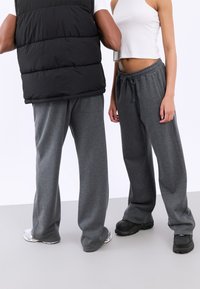 Två personer klädda i lösa grå träningsoveraller och sneakers, en i en vit crop top, den andra i en svart vadderad väst och vit skjorta.