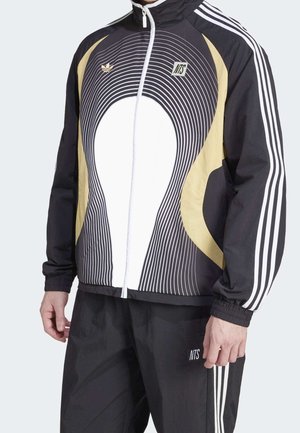 Czarna i beżowa bluza dresowa z białym wzorem z koncentrycznych linii, białym zamkiem błyskawicznym, paskami Adidasa na rękawach oraz logo NTS, noszona z dopasowanymi spodniami.