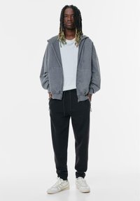 Hanorac gri cu fermoar și buzunare laterale, asortat cu pantaloni de sport negri și adidași albi. Ținuta are un croi lejer și un design casual.