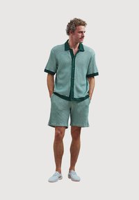 Green short-sleeve shirt and matching shorts with textured fabric and dark green trim. Paired with light blue shoes. Casual fit and design.
