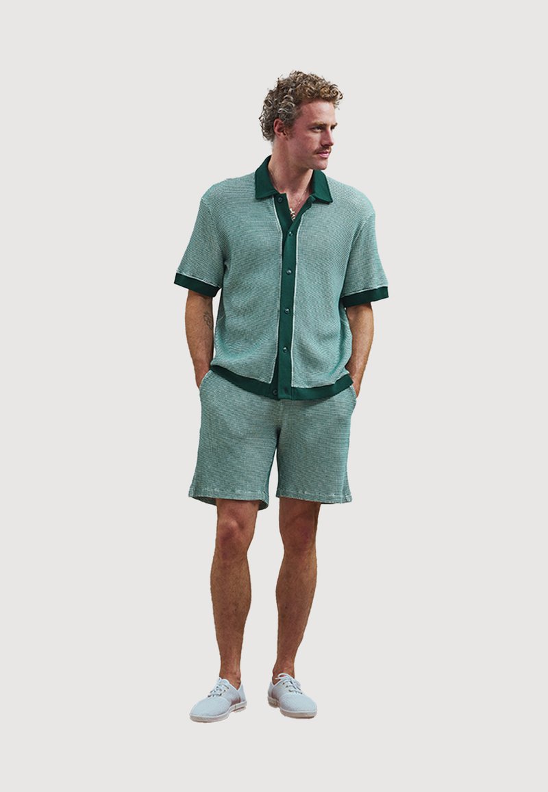 Green short-sleeve shirt and matching shorts with textured fabric and dark green trim. Paired with light blue shoes. Casual fit and design.