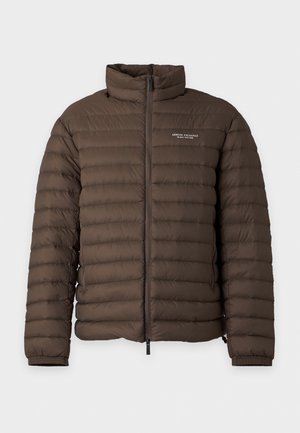 Homme portant une veste matelassée marron, une chemise blanche, un jean bleu et des chaussures noires, debout devant un fond clair uni.