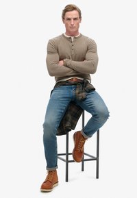 Langærmet beige henley skjorte med knapper, blå jeans, brune støvler og en grønternet skjorte bundet om hoften, siddende på en sort skammel.
