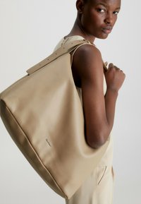 Beige leren tote bag met een zachte, gestructureerde afwerking en een platte, brede strap met verstelbare gaatjes. De merknaam is op de zijkant van de tas gestempeld.