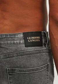 Pantalones vaqueros de mezclilla gris con un bolsillo trasero y una etiqueta negra con la inscripción "GLORIOUS GANGSTA" en letras doradas. Tejido texturizado con detalles de costura.