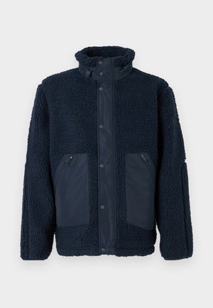 Marineblauwe fleece jas met hoge kraag, drukknoopsluiting, twee ritszakken aan de voorkant en contrasterende stofpanelen op de zakken en de sluiting aan de voorkant.