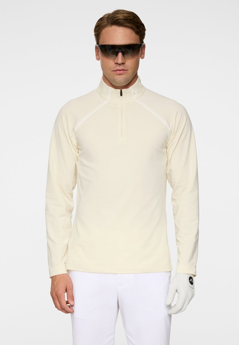 Homme portant une chemise crème à manches longues avec fermeture éclair, pantalon blanc, gant blanc à la main droite et lunettes de soleil sportives foncées, debout devant un fond uni.