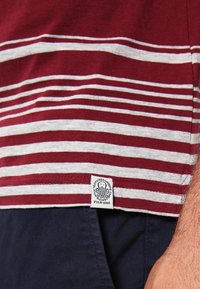 Ourlet de chemise rayé marron et blanc avec étiquette "Pier One" porté sur un pantalon bleu marine foncé, montrant une partie du bras d'une personne.