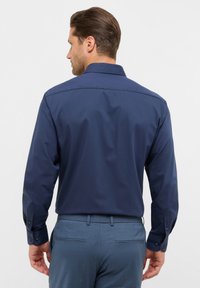 Camicia a maniche lunghe blu navy con colletto aperto, texture liscia e polsini abbottonati, abbinata a pantaloni azzurri chiari.