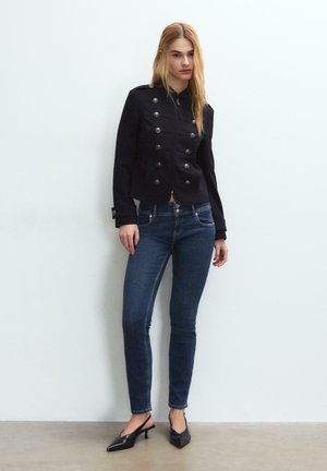 Donna in piedi contro una parete bianca, indossa una giacca nera con bottoni, jeans skinny blu scuro e décolleté nere con punta appuntita.