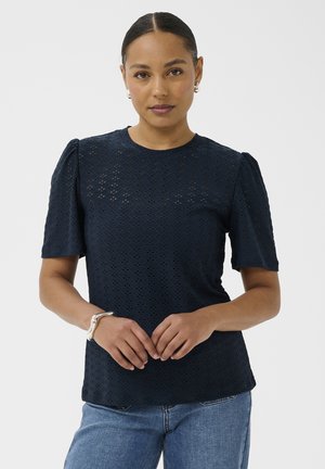 Femme aux cheveux foncés coiffés en chignon, portant un haut à manches courtes bleu marine à motifs et un jean bleu, les mains jointes devant elle.