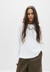 White long-sleeve shirt made from lightweight fabric, with a relaxed fit and round neckline, paired with brown pants featuring side zippers.