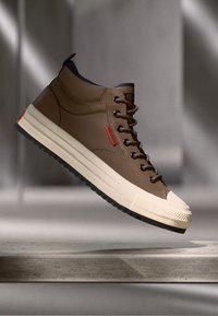 Brown high-top sneaker with a smooth leather upper, cream rubber sole, black accents, and red branding details, featuring metallic eyelets.