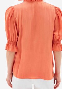 Blouse couleur pêche à manches courtes bouffantes et poignets froncés, avec des rayures verticales et un col à volants. Texture lisse, tissu léger.