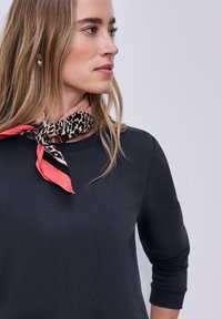 Femme aux cheveux blonds portant une chemise noire à manches longues et une écharpe à motifs corail et noir nouée autour du cou, regardant sur le côté.