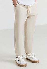 Beige Jogginghosen haben eine weiche Textur, einen verstellbaren Kordelzugbund, Seitentaschen und schmal zulaufende Beine, kombiniert mit weißen Sneakers mit braunen Akzenten.
