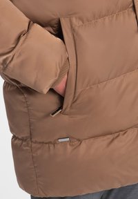 Giacca marrone imbottita con una texture morbida, polsini a coste, tasca laterale con zip e orlo leggermente curvato per una vestibilità comoda.