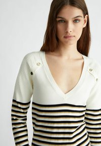 Maglione a manica lunga color crema con scollo a V profondo. Presenta una texture a coste, bottoni dorati accentati e strisce orizzontali nere e dorate.
