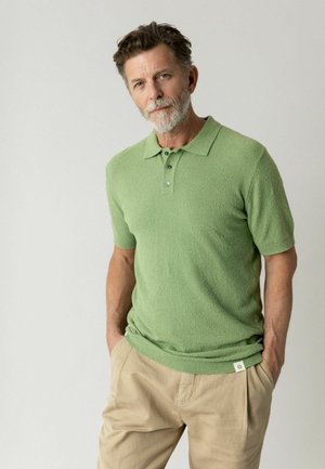Keskeasvanud mees halli habemega, kandes rohelist lühikeste varrukatega polo T-särki ja beeže pükse, seisab käed taskutes hele taust.
