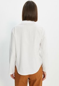 Persona con pelo marrón lacio hasta los hombros, vistiendo una camiseta blanca de manga larga y pantalones marrones, vista desde atrás contra un fondo blanco.