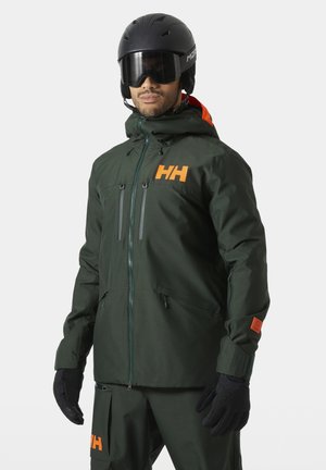 Mand iført mørkegrøn skijakke og bukser med orange HH-logoer, sorte handsker og sort hjelm med tonet visir mod grå baggrund.
