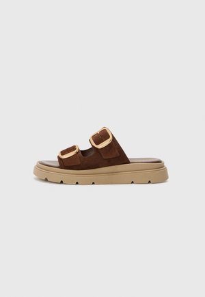 Brown suede slide sandal with two adjustable straps and gold-tone buckles. Thick beige sole with a textured grip pattern.
