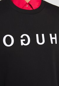 Un sweat-shirt noir en tissu doux avec des lettres blanches audacieuses épelant "HUGO", avec une chemise colorée rouge visible en dessous.