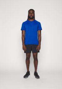 Blaues kurzärmliges Sportshirt, schwarze Shorts und schwarze Laufschuhe. Glatter Stoff, figurbetontes Design und minimalistischer Stil.