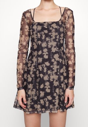 Sie trägt ein schwarzes, transparentes langärmliges Kleid mit beigen Blumenmustern und einem quadratischen Ausschnitt über einem schwarzen Trägertop.