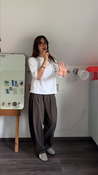 Camicia bianca, ampia; pantaloni marroni a gamba larga; scarpe grigie. In piedi davanti a uno specchio. Decorazioni da parete visibili sullo sfondo.