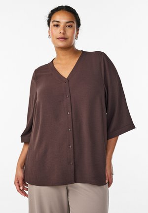 Ženska, oblečena v široko temno rjavo srajco na gumbe s krilnimi rokavi dolžine tri četrtine in bež hlače, stoji pred belim ozadjem.