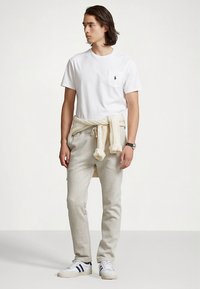 Λευκό βαμβακερό μπλουζάκι με τσέπη στο στήθος, μπεζ φόρμες και κρεμ πουλόβερ δεμένο γύρω από τη μέση. Λευκά αθλητικά παπούτσια με σκοτεινές ρίγες.