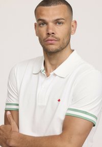 Polo shirt blanche avec des manches à rayures vertes, arborant un logo en forme de diamant rouge sur la poitrine. Texture lisse et design de col classique.