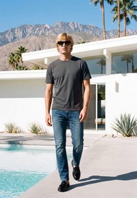 Junger Mann mit Sonnenbrille, schwarzem T-Shirt und Jeans geht an einem Pool mit modernem Haus, Palmen und Bergen im Hintergrund vorbei.