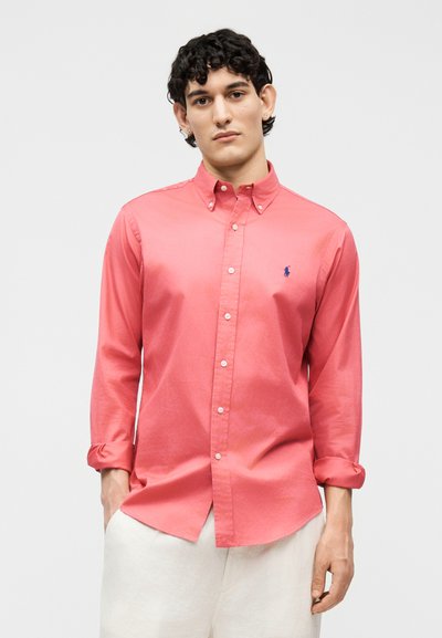 Νεαρός άνδρας με κοντά σγουρά μαλλιά, φοράει ένα ροζ κοραλλί πουκάμισο με κουμπιά και μανίκια που έχουν γυριστεί, καθώς και ανοιχτό μπεζ παντελόνι, στέκεται μπροστά σε μια απλή φόντο.