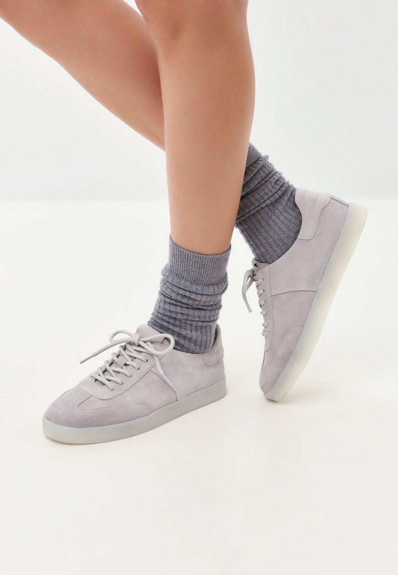Jambes portant des chaussettes côtelées grises et des baskets en daim gris clair avec des semelles blanches sur fond blanc.
