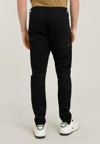 Pantalones de denim negros con corte slim, que cuentan con bolsillos traseros y detalles de costura sutiles. Combinados con zapatillas blancas y una camiseta color tostado.