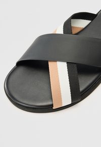 Svart slide-sandal med ett slätt, matt rem över en texturerad sula. Accentuerad med ränder i persika, vitt och svart.