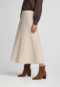 Kvinde iført en beige A-line midi-nederdel, brun bluse med lange ærmer og brune ruskind ankelstøvler med blokhæle, stående foran en lys baggrund.