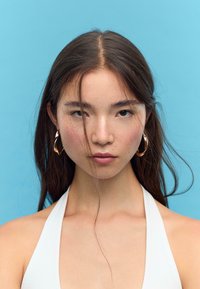 Model med langt, mørkt hår, iført en hvid halterneck top. Bemærkelsesværdige guld hoops, glat hud med lette fregner og neutral baggrund.