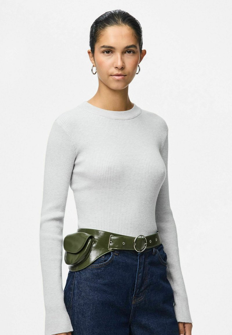 Šedý pruhovaný top s dlouhým rukávem spárovaný se zeleným lesklým páskem s kulatou sponou a kapsičkou, nošený s modrými džínovými kalhotami.