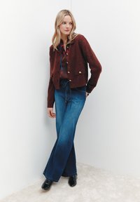 Blonde Frau, die eine bordeauxfarbene geknöpfte Strickjacke, eine bedruckte Bluse, hochgeschnittene weit geschnittene Jeans und schwarze Stiefeletten trägt, steht vor weißen Wänden.