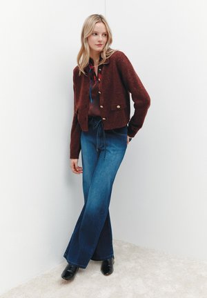 Blonde Frau, die eine bordeauxfarbene geknöpfte Strickjacke, eine bedruckte Bluse, hochgeschnittene weit geschnittene Jeans und schwarze Stiefeletten trägt, steht vor weißen Wänden.