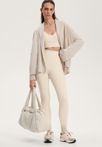 Beige avointa neuletakki, jonka päällä on yhteensopiva lyhyt toppi ja korkea vyötäröiset leggingsit. Kantaa pehmeää, tikattua olkalaukkua. Kengät täydentävät asua.