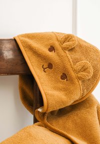 Dětská osuška s kapucí z měkké, strukturované látky zlatavě žluté barvy. Má uši medvěda, hnědě vyšívané oči a pohodlný design.