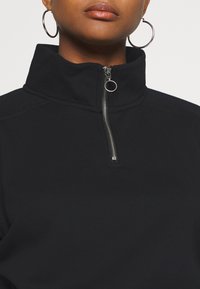 Svart tröja med högkrage och halvdragkedja, med en slät yta och en silverfärgad dragkedja som accent.