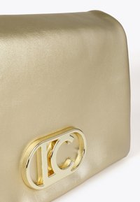 Zlatá metalická kabelka na ruku s výrazným zlatým logem, která se vyznačuje hladkým povrchem a elegantním designem.